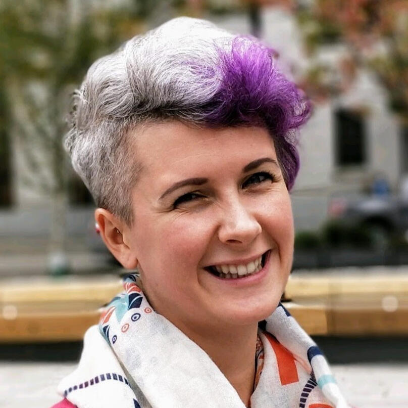 A person faces the camera smiling. Their hair is gray and has a purple streak.